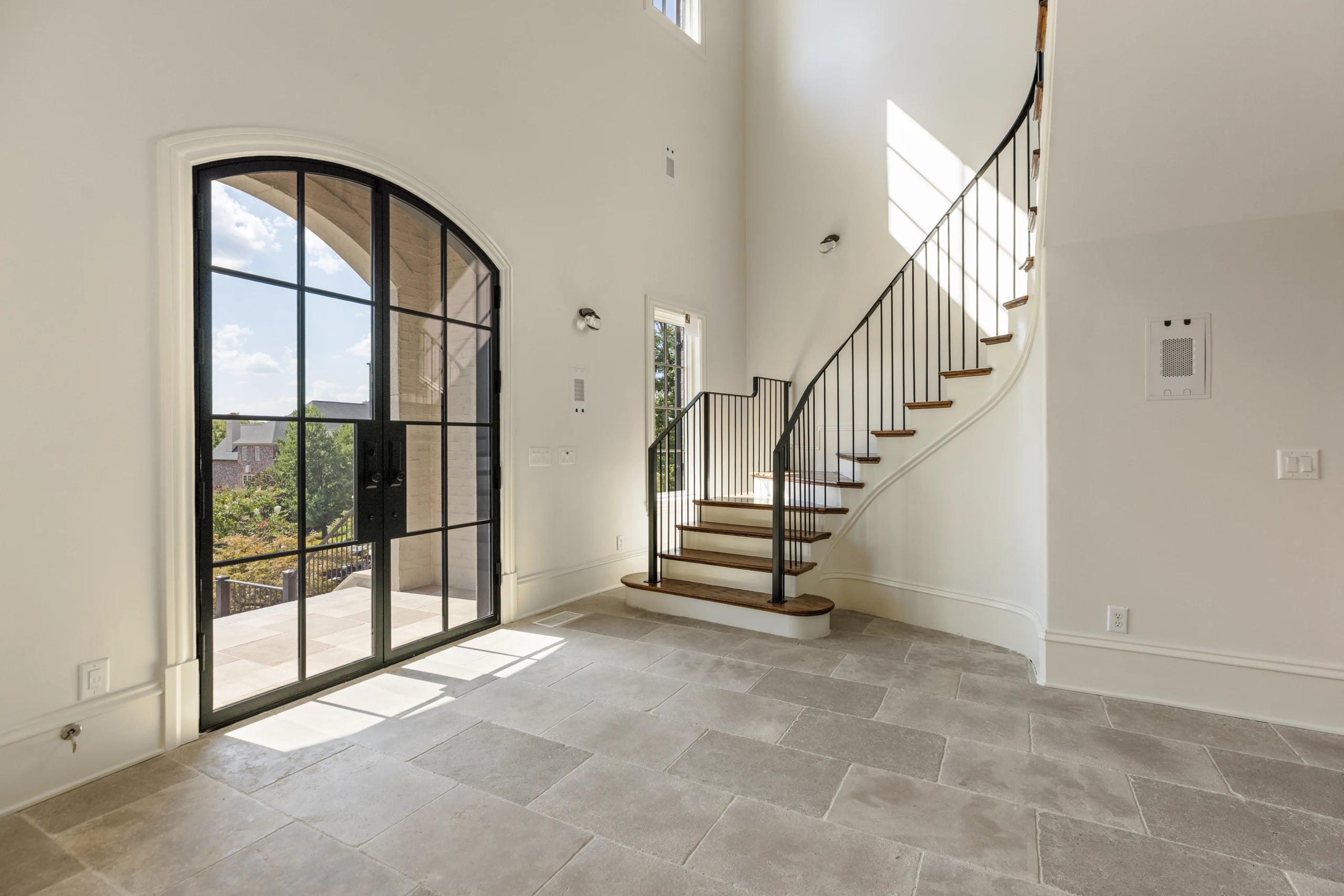 Arched iron entry door beside a curved staircase, with pale limestone flooring